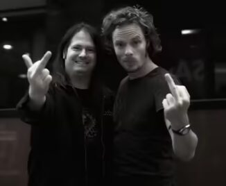 Two men facing the camera, both showing middle fingers. One has long hair and wears a jacket, the other has short hair and wears a watch. Black and white photo.